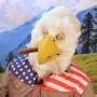 Man wearing latex prosthetic beak and makeup with feathers to look like an anthropomorphic American-themed bald eagle with cigar.