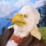 Man wearing latex prosthetic beak and makeup with feathers to look like an anthropomorphic bald eagle with cigar.