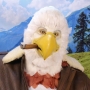 Man wearing latex prosthetic beak and makeup with feathers to look like an anthropomorphic bald eagle with cigar.